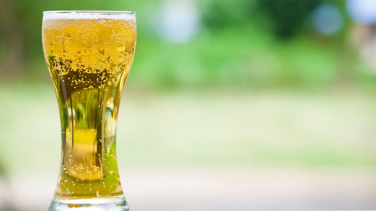 A clear, curvy glass filled with golden, bubbly liquid sits on a surface, with a blurred green and white background suggesting an outdoor setting.