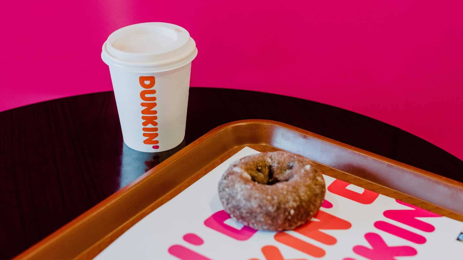 A white Dunkin' cup with a lid and an iced donut covered in sugar sit on a tray atop a table. The background is bright pink.