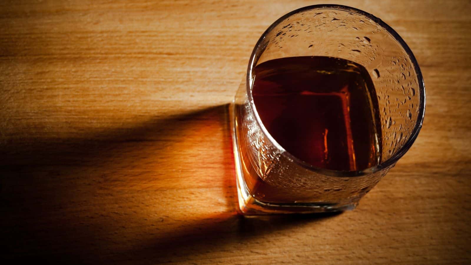A glass filled with a dark brown liquid, possibly whiskey or another spirit, sits on a wooden surface with light casting a shadow to the left.