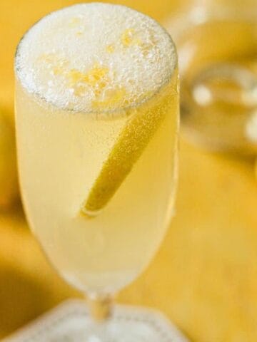 A champagne flute filled with a bubbly, light yellow cocktail garnished with a lemon twist. In the blurred background, there are several lemons, some whole and some sliced. The setting has a warm, golden tone.