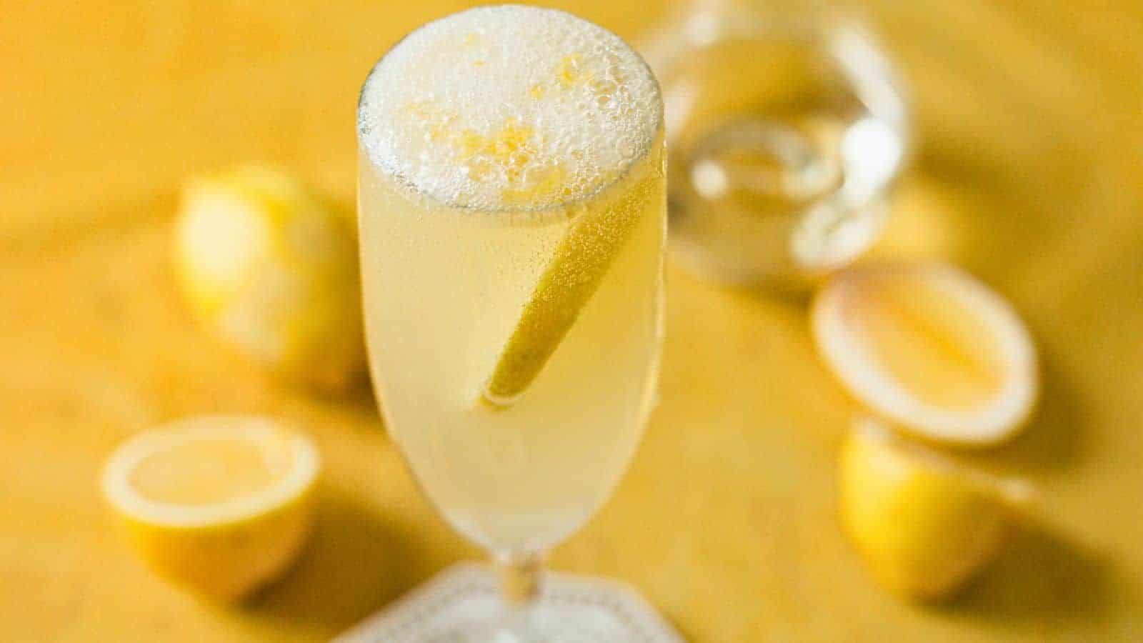 A champagne flute filled with a bubbly, light yellow cocktail garnished with a lemon twist. In the blurred background, there are several lemons, some whole and some sliced. The setting has a warm, golden tone.