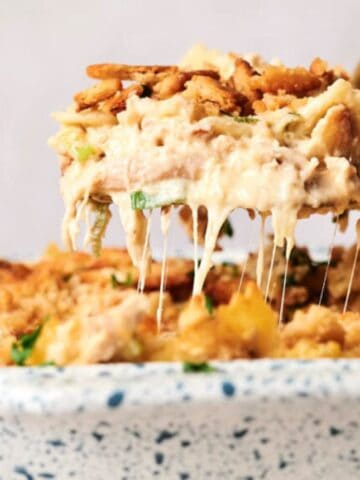 A serving of a cheesy casserole with a crispy topping is being lifted from a speckled baking dish. The dish contains layers of shredded cheese, creamy sauce, and herbs. A wooden spoon is in use.