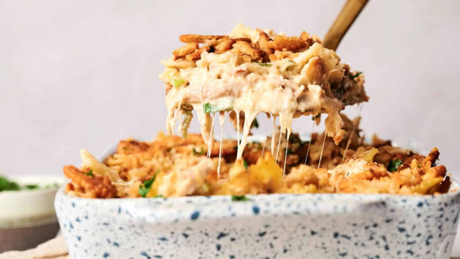 A serving of a cheesy casserole with a crispy topping is being lifted from a speckled baking dish. The dish contains layers of shredded cheese, creamy sauce, and herbs. A wooden spoon is in use.