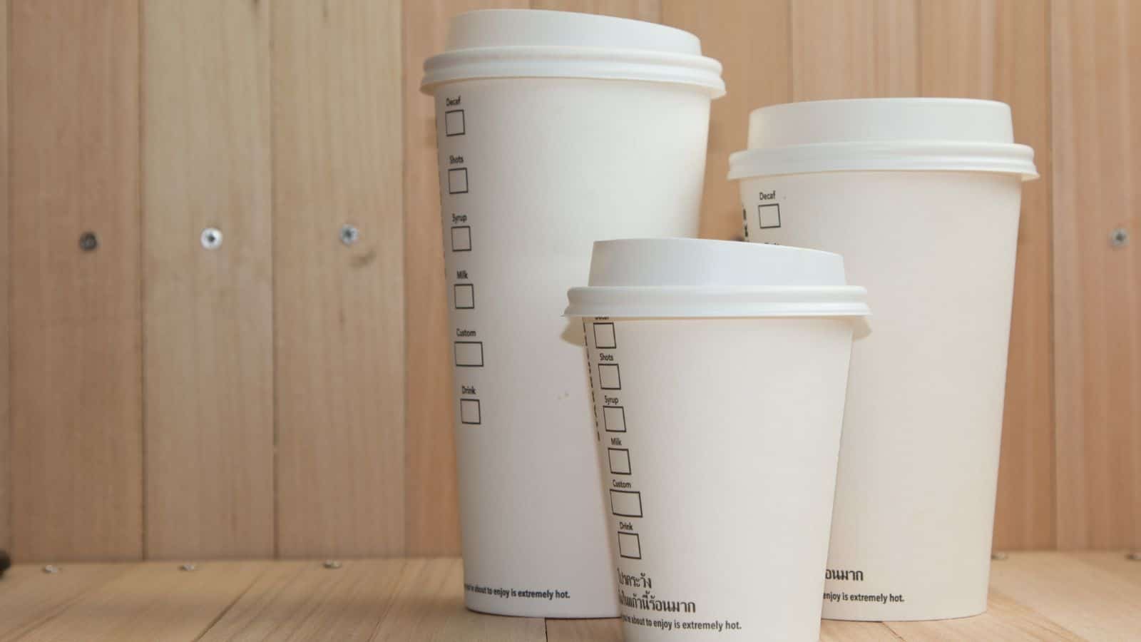 Three white disposable coffee cups with lids are displayed on a wooden surface. The cups vary in size from small to large. Each cup has a series of checkboxes on the side for customization. The background features light wooden paneling.