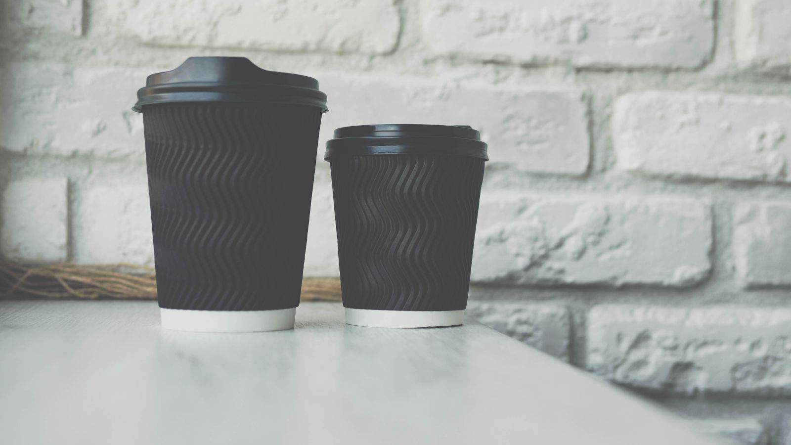 Two black disposable coffee cups with lids are placed on a light-colored table. The larger cup is on the left, and the smaller one on the right. The background features a white brick wall.