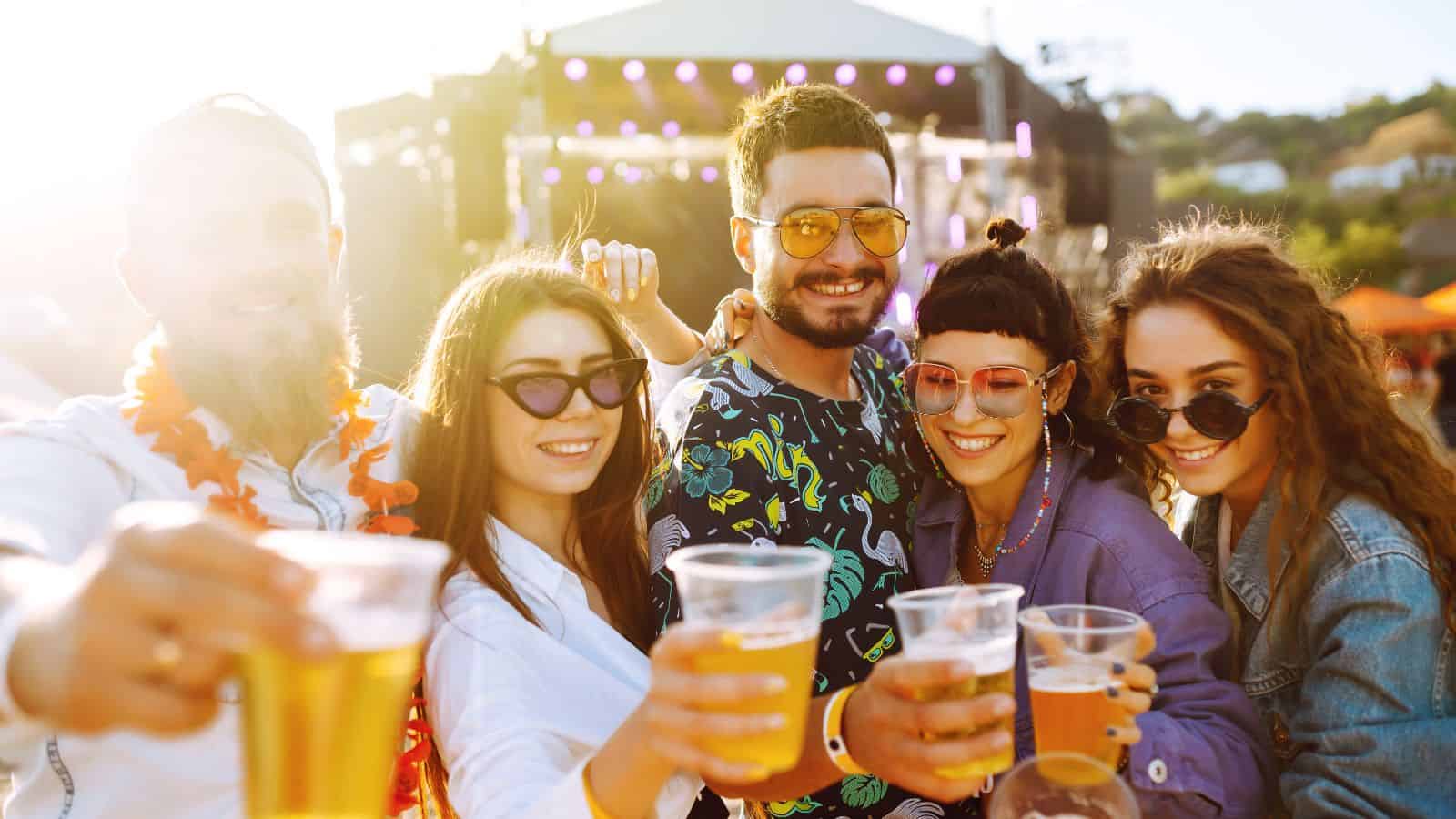 Five people wearing sunglasses and casual clothes smile and hold plastic cups of beer toward the camera at an outdoor event with a stage visible in the background. The scene is brightly lit by sunlight.