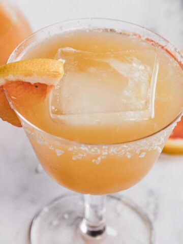 A cocktail in a glass with a salted rim, large ice cube, and a grapefruit wedge garnish. A grapefruit slice, a lime wedge, and a carafe with an orange drink are in the background on a marble surface.
