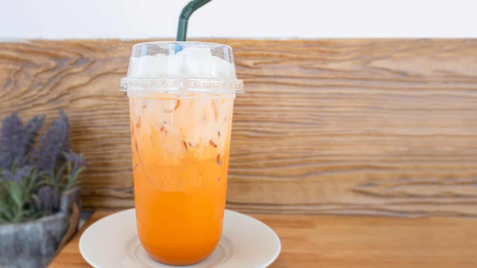 A clear plastic cup filled with iced orange-colored tea with milk, covered with a plastic lid and a green straw, sits on a white saucer against a wooden background. A small potted plant is partially visible to the left.