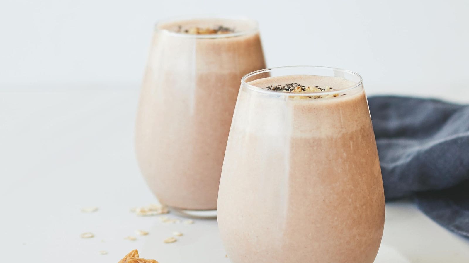 Two glasses filled with a creamy brown smoothie are placed on a light surface. The smoothies are slightly frothy on top and have small chia seeds sprinkled on them. A dark cloth napkin is partially visible in the background.