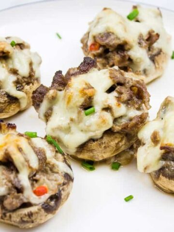 Five stuffed mushrooms topped with melted cheese and garnished with chopped chives are arranged on a white plate with a thin dark border.