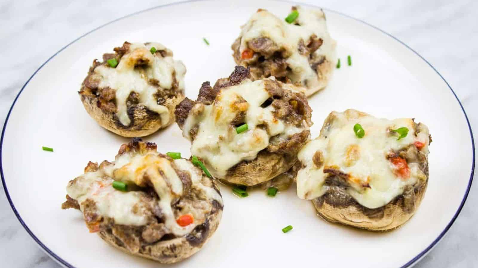 Five stuffed mushrooms topped with melted cheese and garnished with chopped chives are arranged on a white plate with a thin dark border.