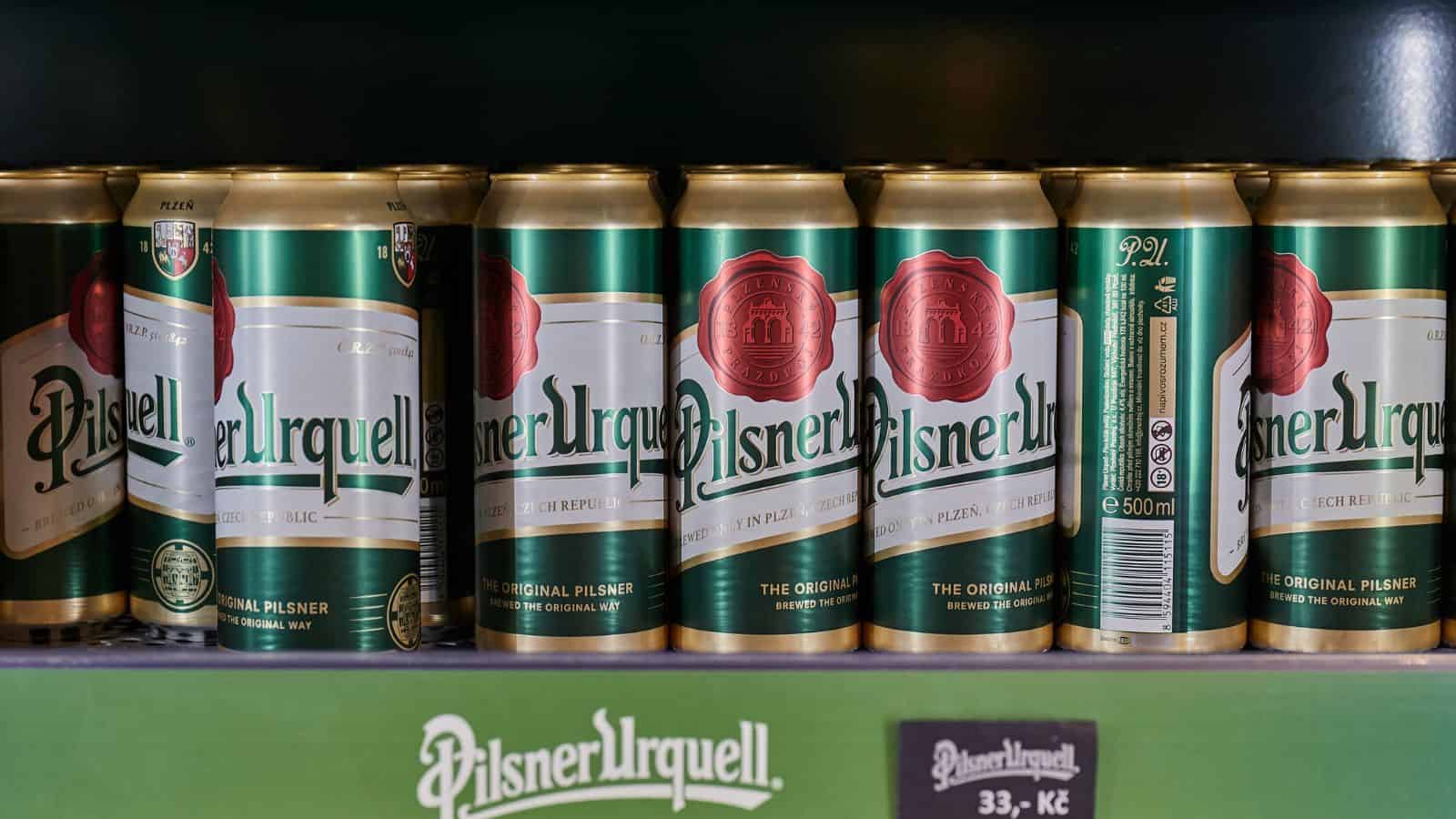A row of Pilsner Urquell beer cans is displayed on a store shelf. The cans are predominantly green with red and white labels, featuring the brand name prominently. A sign below shows the price and additional information in another language.