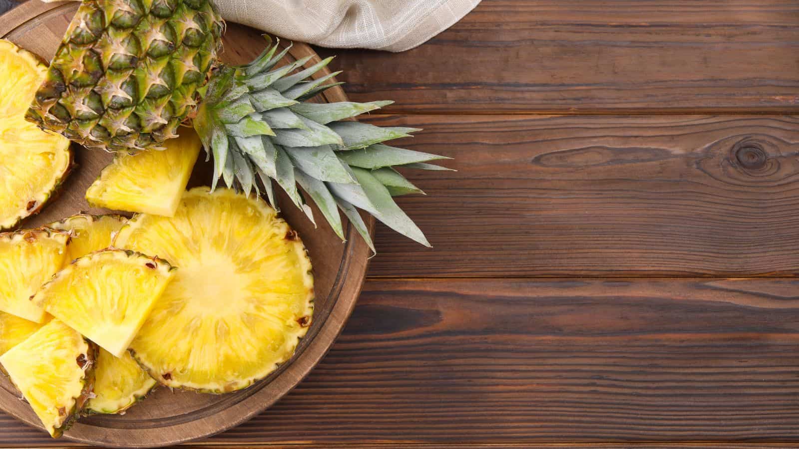 A wooden tray holds a whole pineapple with its leafy top, along with several round and triangular slices of fresh pineapple. The tray is placed on a wooden surface with a beige cloth in the background.