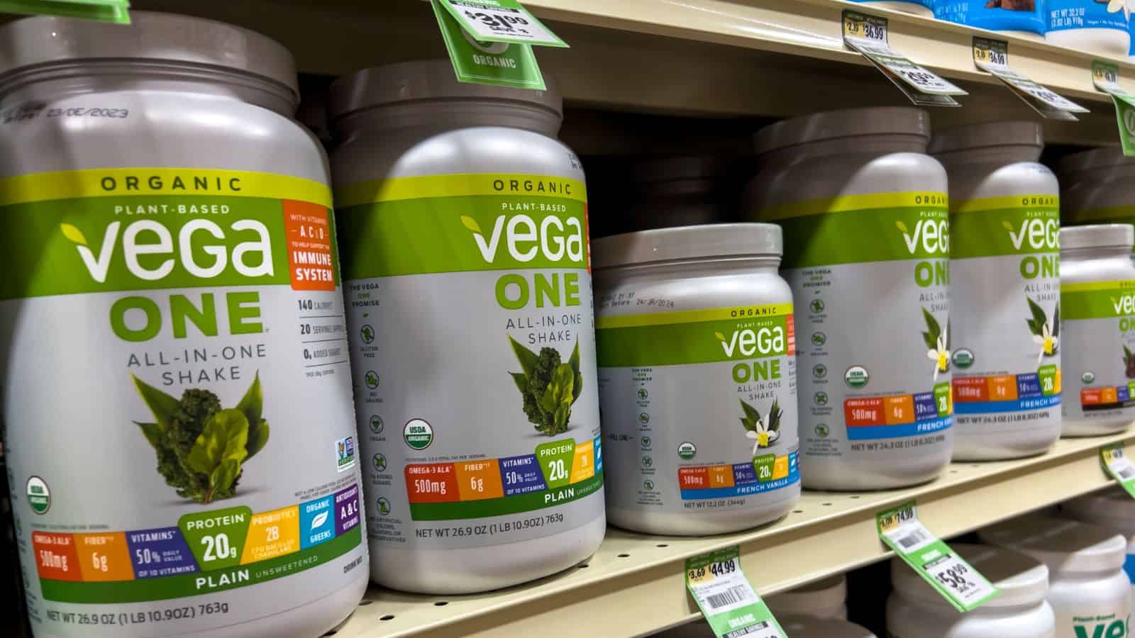 Several containers of Vega One Organic Plant-Based All-In-One Shake in plain flavor are displayed on a grocery store shelf, each container showing nutrition information and a green label. Price tags are visible above the products.