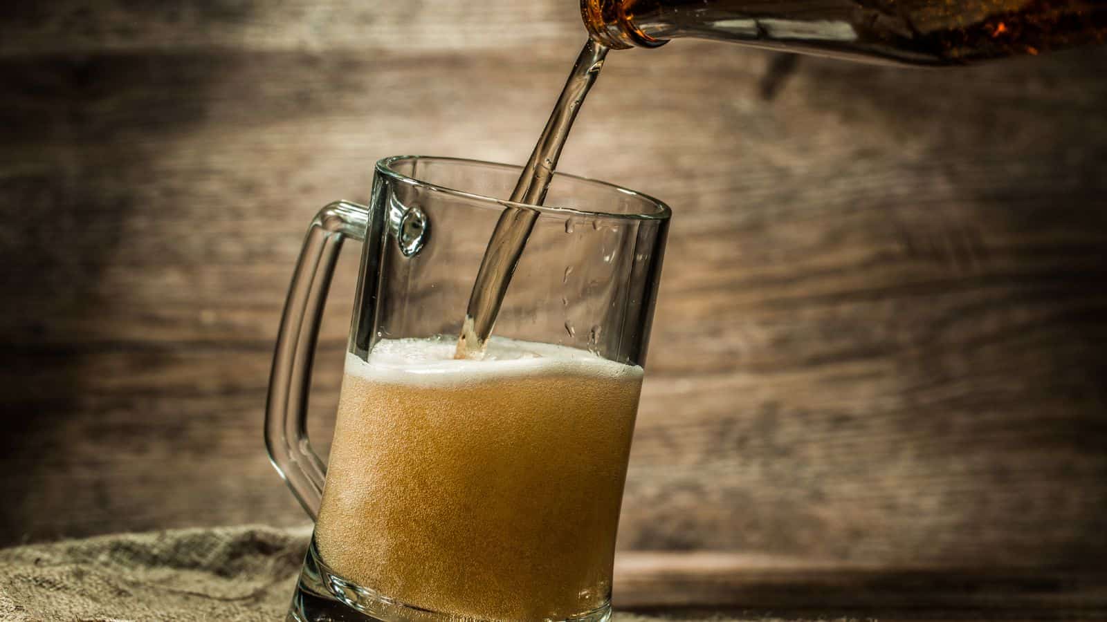 A glass mug is being filled with beer from a bottle. The beer is golden with a foamy head forming. The background is wooden, giving a rustic feel to the setting.