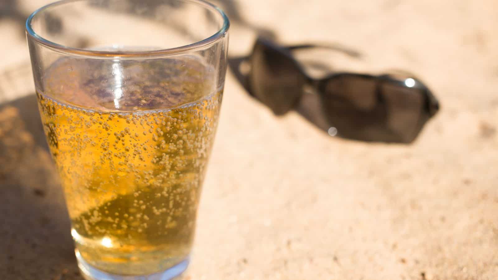 A clear glass of a fizzy, golden-colored beverage sits on a sandy surface. In the background, a pair of dark sunglasses rests on the sand, slightly out of focus.