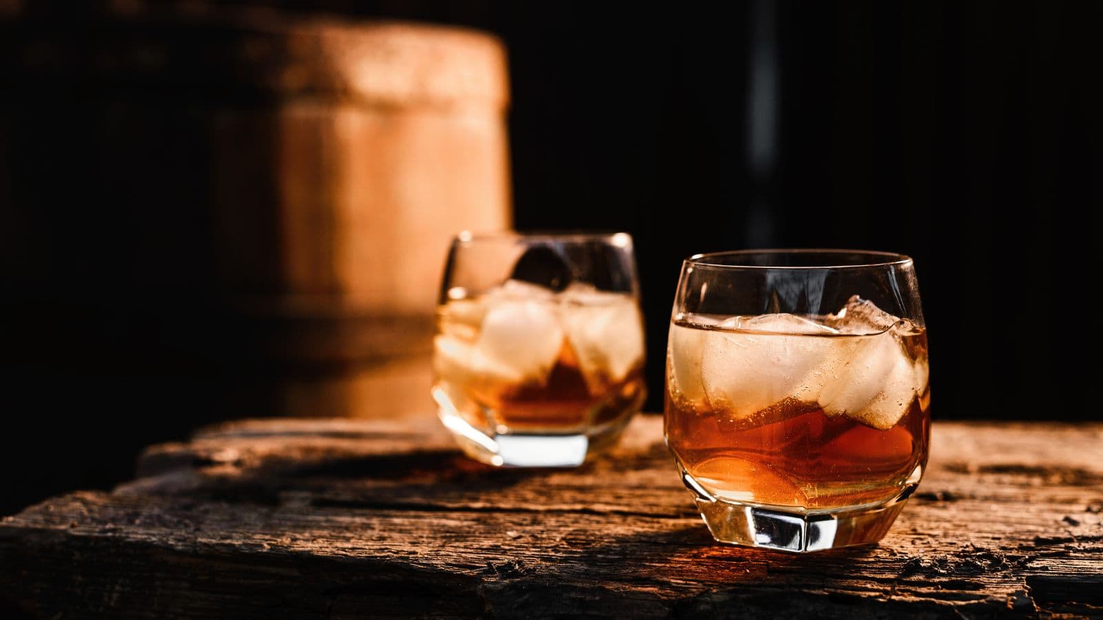 Two glasses of whiskey with ice cubes sit on a rustic wooden surface, with a blurred barrel in the background. The lighting is warm and highlights the amber color of the drinks.