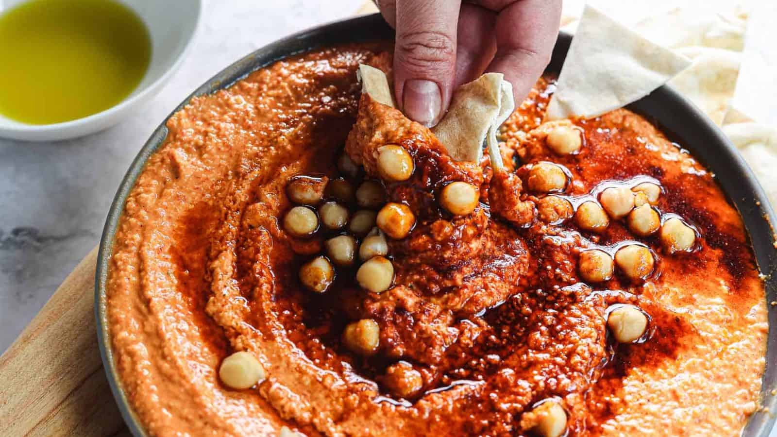 A bowl of creamy hummus topped with chickpeas and a red sauce, placed on a wooden board. Pita bread pieces and a small bowl of olive oil are nearby on a marble surface.