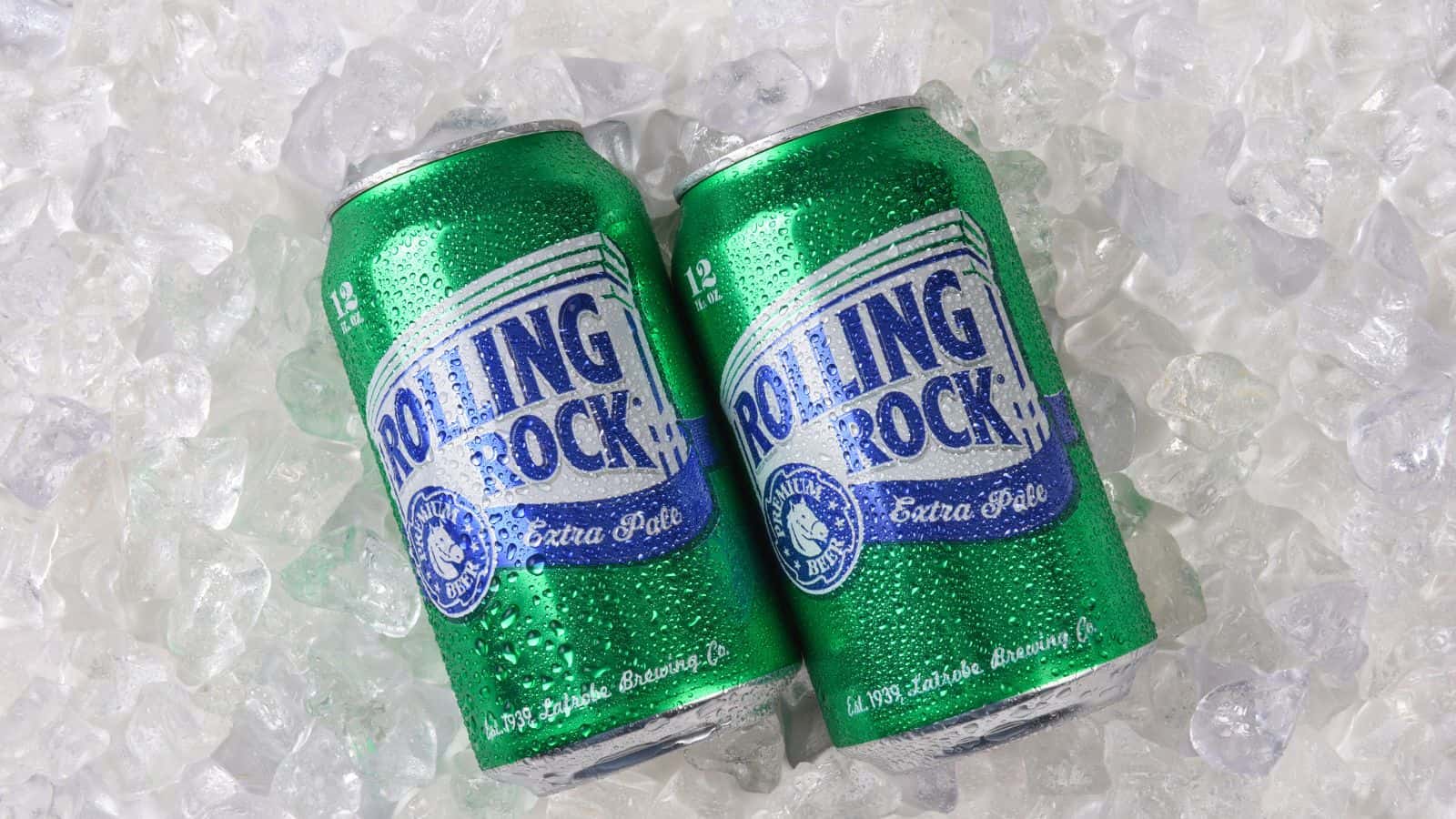 Two green Rolling Rock beer cans with condensation are lying on a bed of ice cubes. The cans have blue and white labels displaying the brand name and logo.