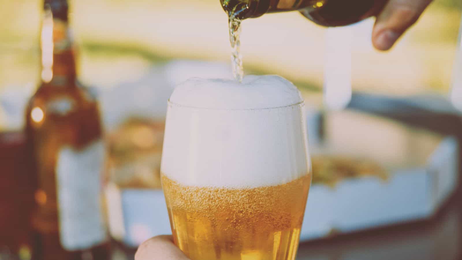 A glass is being filled with beer, creating a large foam head. Another bottle and a pizza box are visible in the blurred background on a table outdoors.