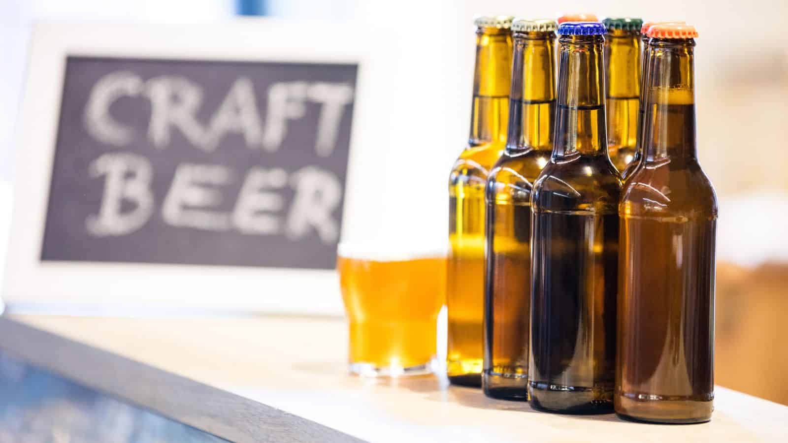 Five brown glass beer bottles, each with a different colored cap, are lined up on a counter next to a glass of beer. A sign in the background reads &ldquo;CRAFT BEER&rdquo; in large white letters on a chalkboard.