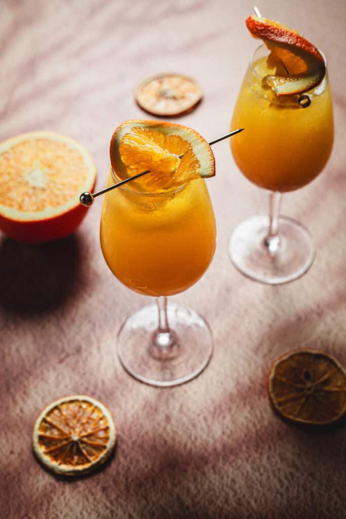Two wine glasses filled with an orange-colored drink, garnished with orange slices on metal picks, are on a table with dried orange slices and a halved fresh orange.