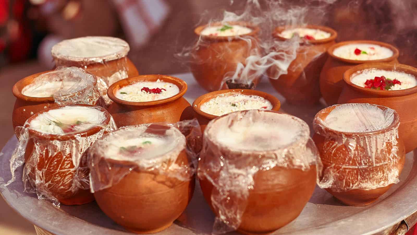 Several clay pots filled with steaming, creamy desserts are arranged on a round surface. Each pot is topped with a clear wrap, and some have garnishes, like red and green pieces. The steam is visible above the pots, indicating warmth.
