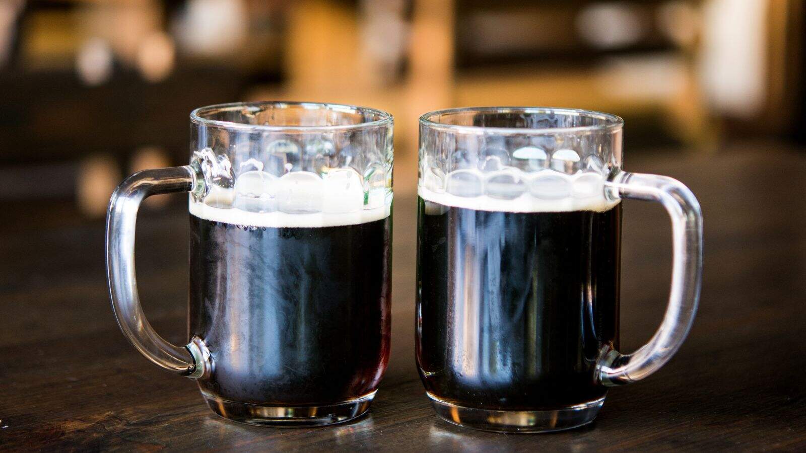 Two glass mugs filled with dark beer are placed side by side on a wooden table. The mugs have clear handles, and a frothy head is visible on the surface of the beer. The background is softly blurred, emphasizing the mugs.