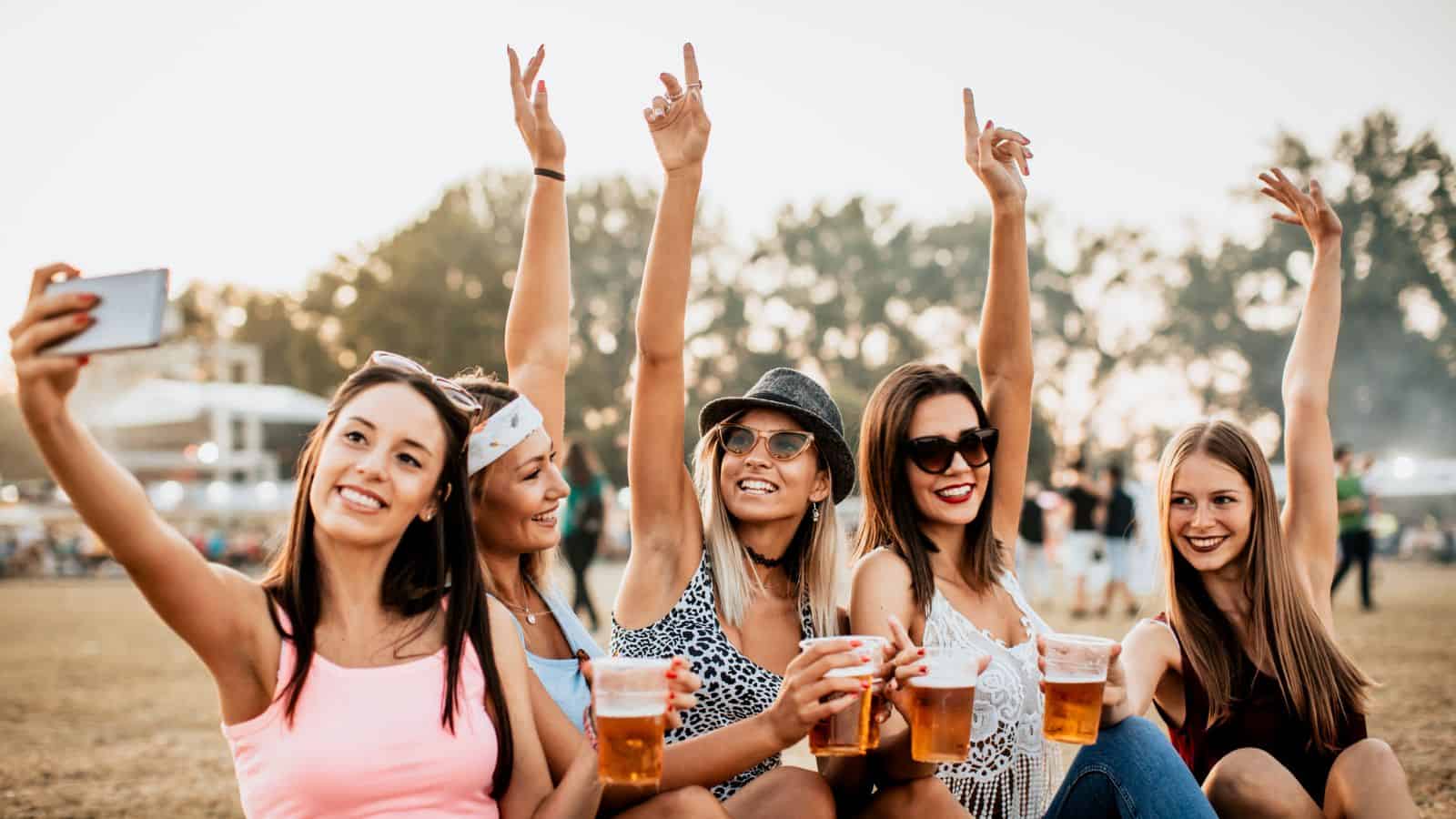 Five young women are sitting outdoors, holding plastic cups of beer, smiling, and posing for a selfie. Some have their arms raised in the air. Trees and festival tents are visible in the background.