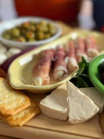 A selection of appetizers on a wooden board, featuring sliced meats, cheese, olives, tomatoes, and crackers with a colorful cookware in the background.