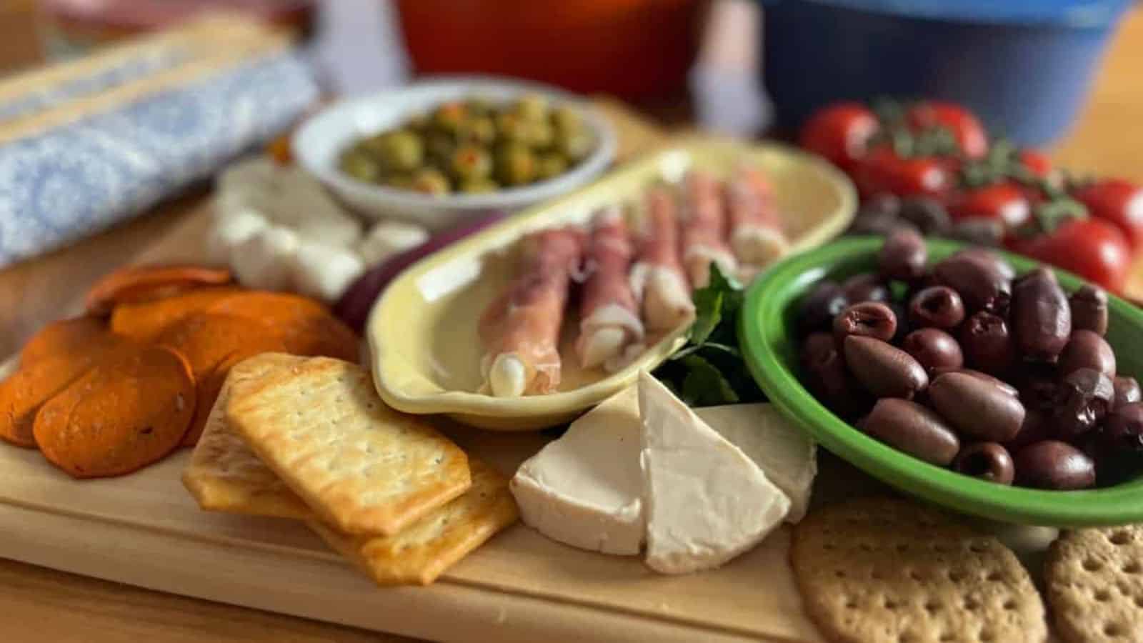 A selection of appetizers on a wooden board, featuring sliced meats, cheese, olives, tomatoes, and crackers with a colorful cookware in the background.