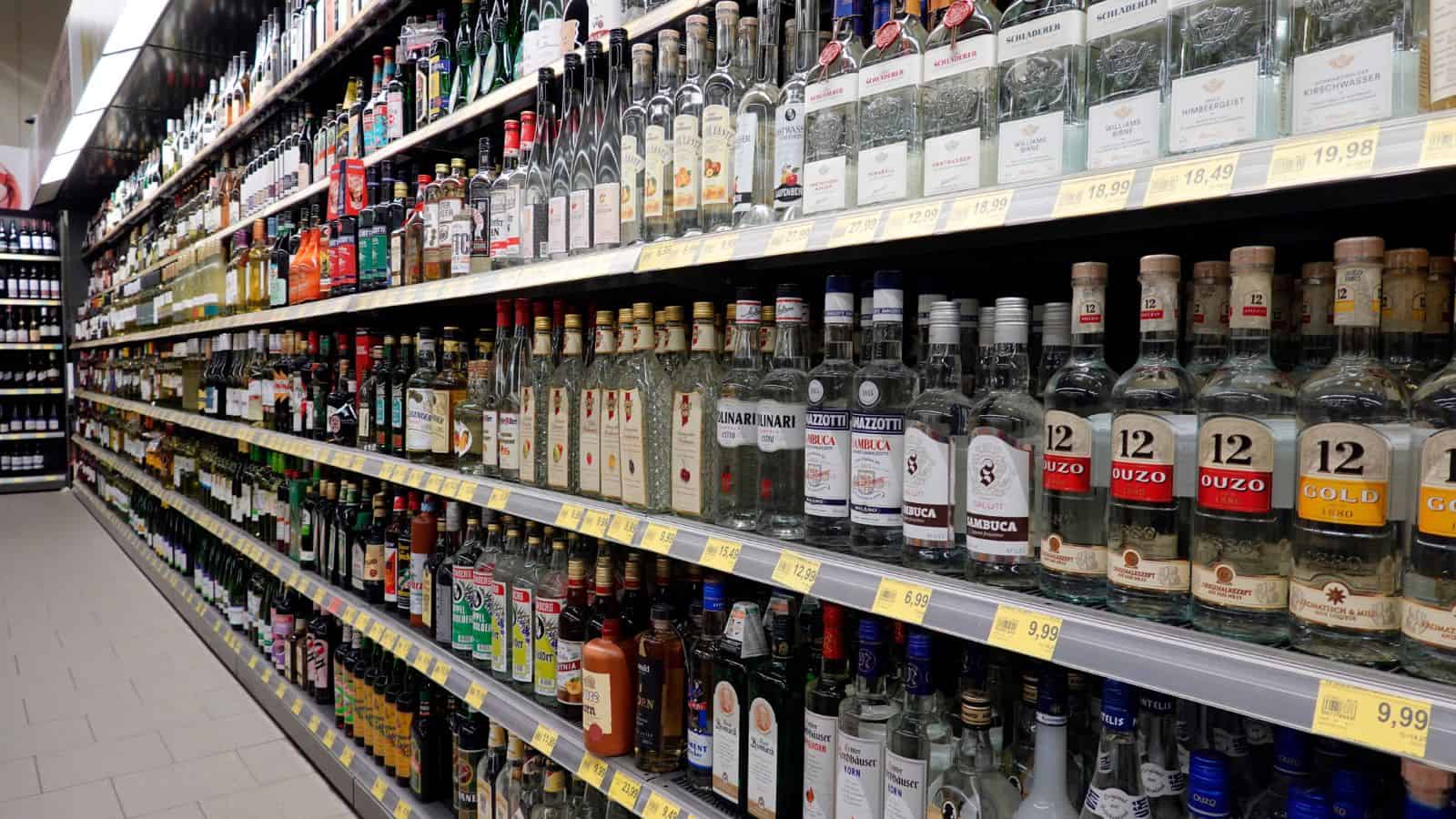 Shelves filled with various bottles of liquor, including vodka, whiskey, and gin, in a store. Price tags are visible below each product. The floor is tiled, and the lighting is bright, highlighting the colorful labels on the bottles.