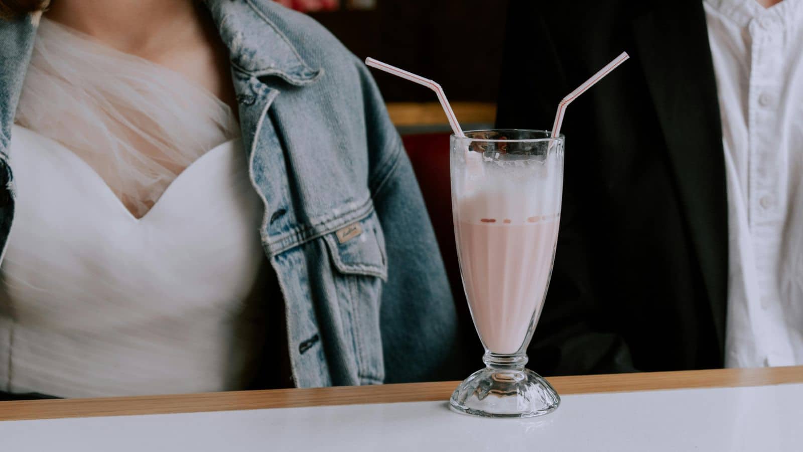 A glass of pink milkshake with two straws sits on a table between two people, one wearing a white dress and denim jacket, and the other in a white shirt and dark jacket.