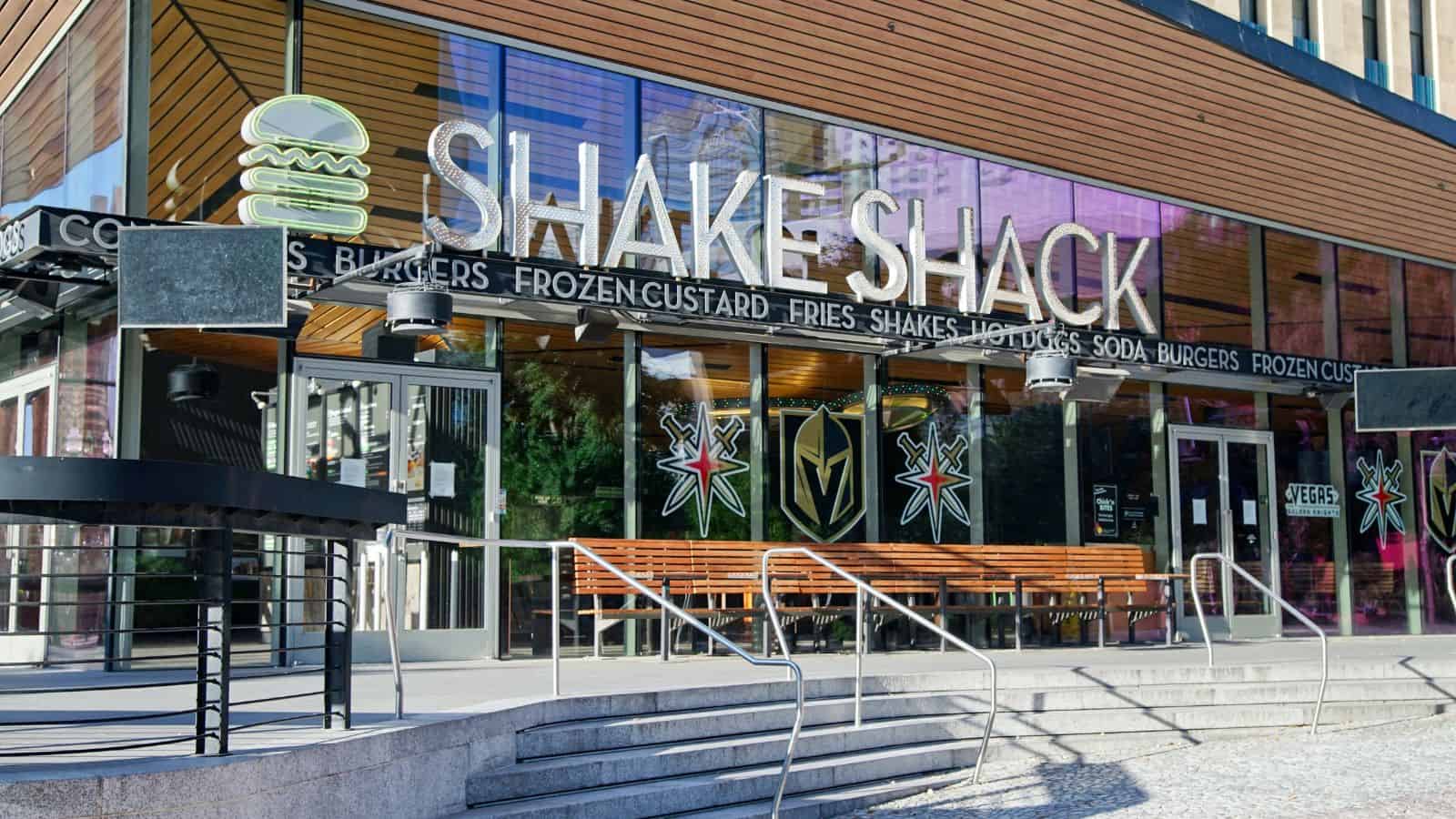 The entrance of a Shake Shack restaurant with large glass windows, a neon burger sign, and “Shake Shack” written above the door; metal railings and steps lead up to the entrance.