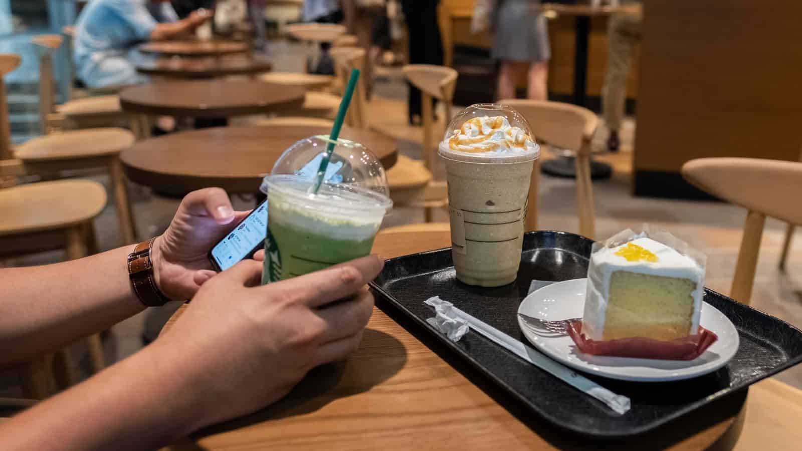 A person holding a green iced drink and a smartphone sits at a wooden table with a tray. The tray has a caramel-topped iced drink and a slice of cake on a plate with a fork and knife. Blurred background shows more tables and chairs.