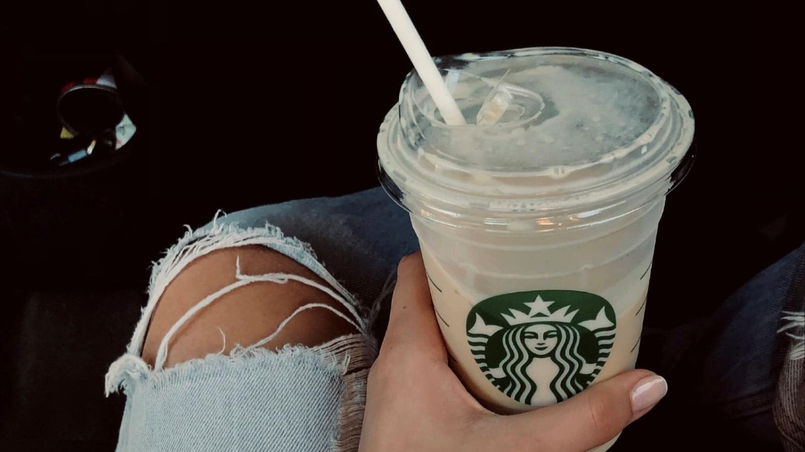 A person holds a clear plastic cup with a Starbucks logo, filled with an iced beverage and a straw. The person is wearing ripped jeans and has light-colored nail polish.