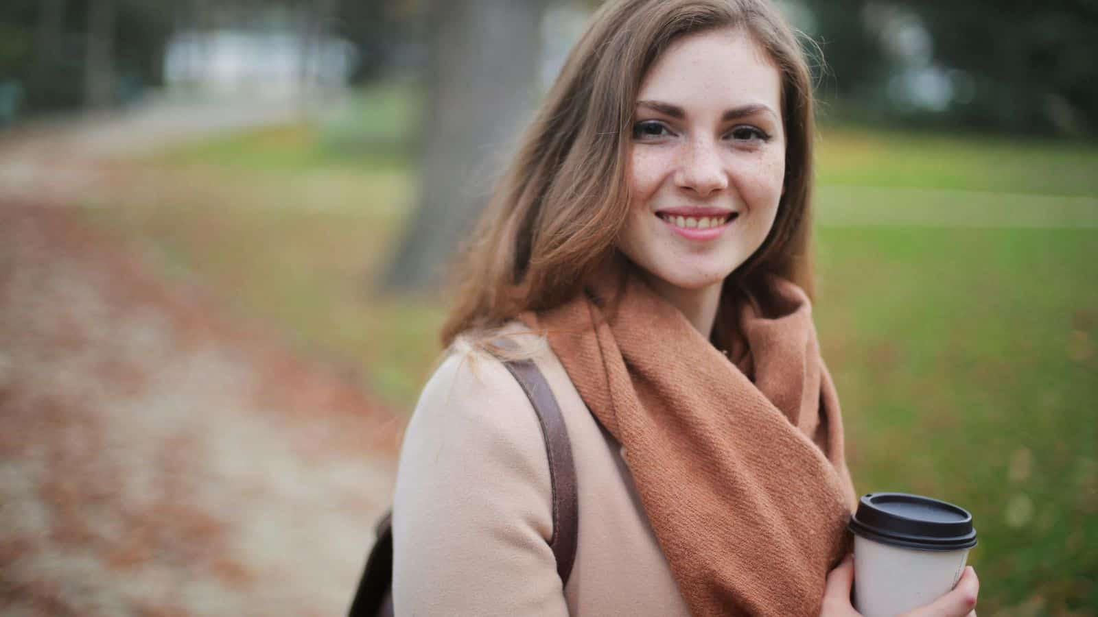 A woman with long hair is smiling while holding a takeaway coffee cup. She is wearing a light-colored coat and brown scarf. The background is a park with autumn leaves on the ground.