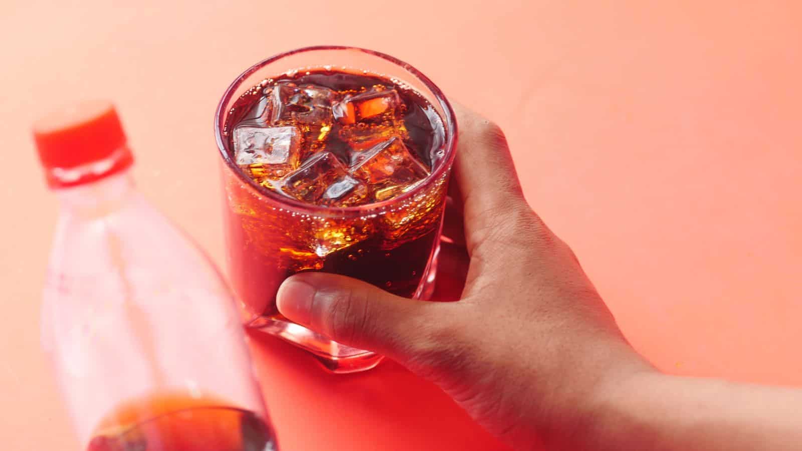 A hand holding a glass filled with a dark, fizzy beverage and ice cubes. A plastic soda bottle with a red cap is partially visible beside the glass. The background is a bright orange surface.