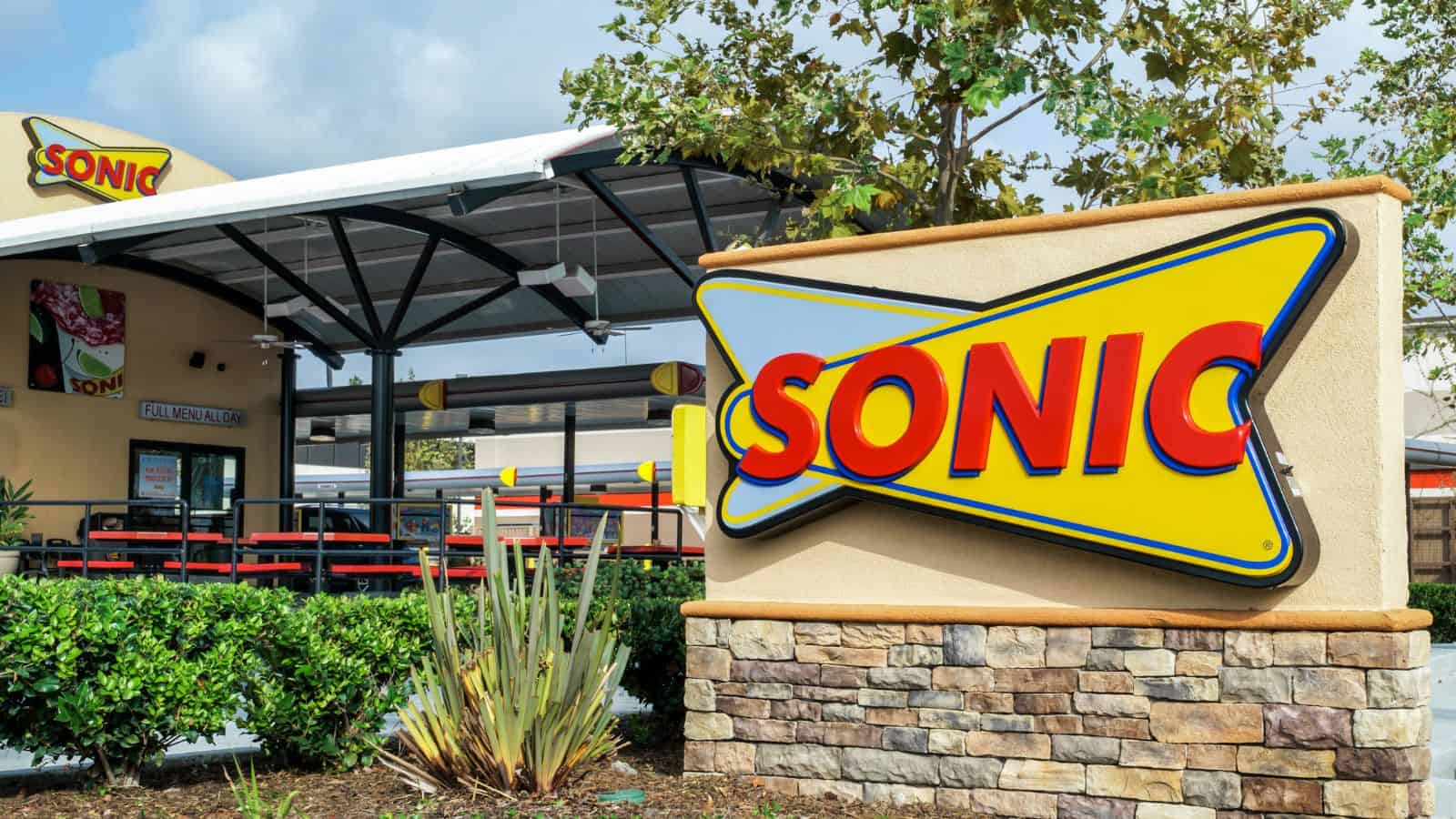 A Sonic Drive-In restaurant exterior featuring a large yellow and red Sonic sign, outdoor seating under a canopy, greenery, and a stone and stucco wall in the foreground.