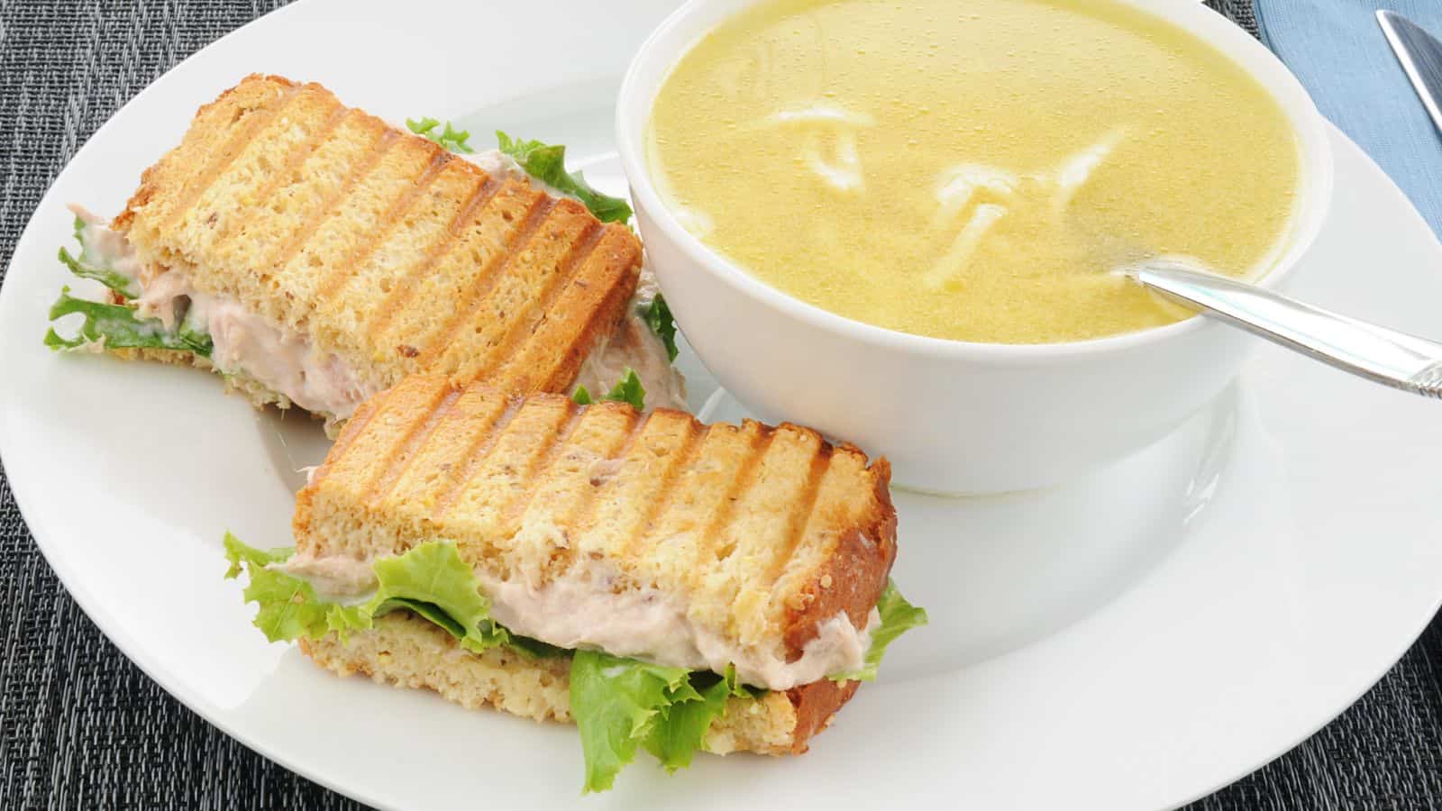 A white plate with a bowl of soup and a spoon, beside a toasted sandwich with lettuce and filling. The soup appears creamy with noodles, and the sandwich has grill marks. The setting is on a textured surface.