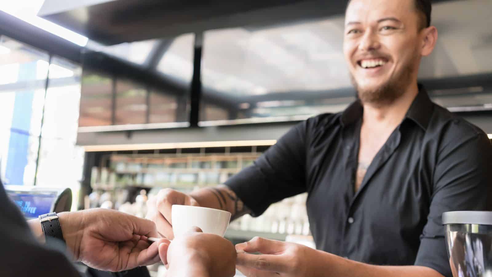 A smiling barista in a black shirt hands a white coffee mug to a customer across a counter in a modern cafe. The background features shelves with various items and large windows.
