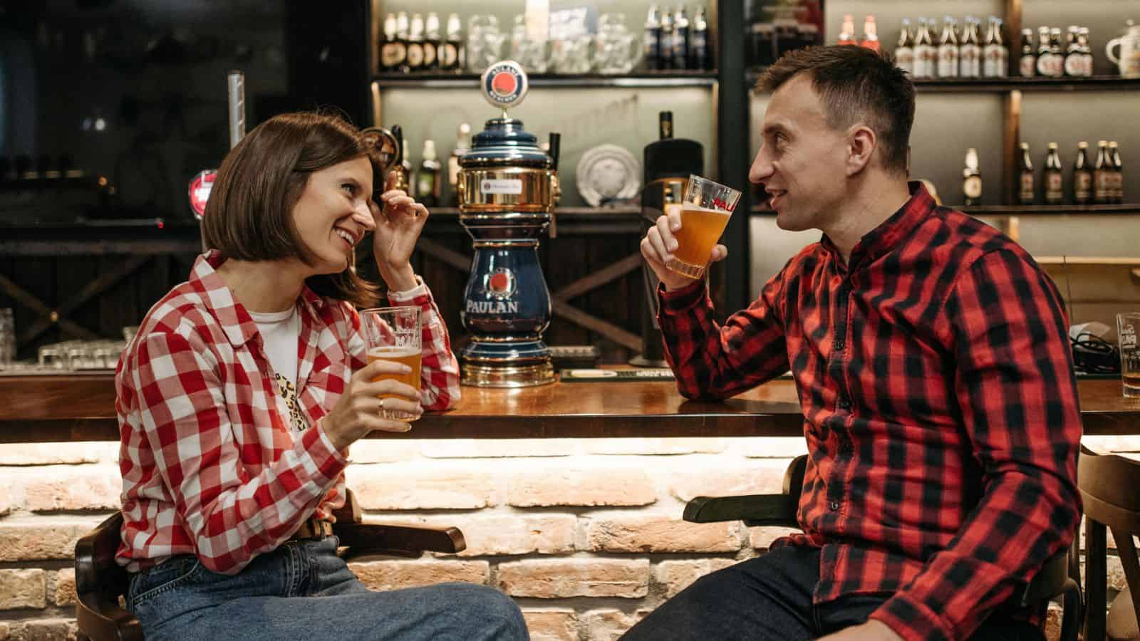 A woman and a man sit at a bar, both holding glasses of beer. They wear plaid shirts and face each other while smiling. In the background, there are various bottles on shelves and a beer tap at the center of the bar.