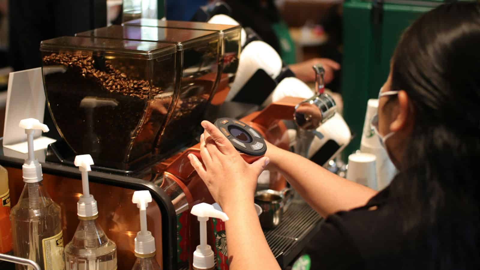 A barista prepares a drink next to a coffee grinder and syrup bottles. The coffee beans are visible in a transparent container. Various nozzles and equipment surround the work area, suggesting a cafe setting.