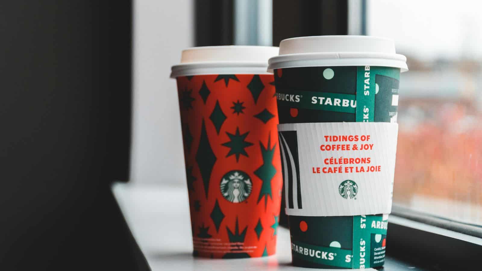 Two Starbucks coffee cups with holiday designs sit on a windowsill. The cup in the foreground is green with a white sleeve and holiday message, while the cup in the background is red with star and tree patterns.