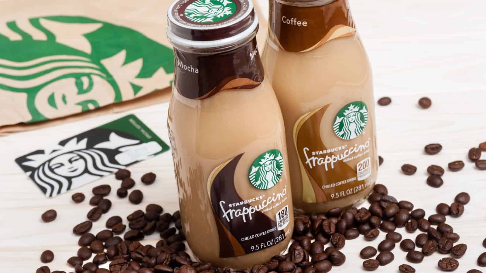Two bottles of Starbucks Frappuccino, one mocha and one coffee flavor, stand on a light surface surrounded by coffee beans, with a Starbucks paper bag and gift card visible in the background.