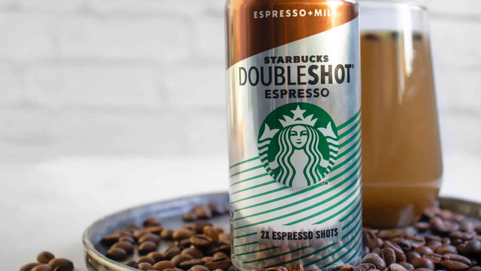 A can of Starbucks Doubleshot Espresso and Milk stands on a plate with coffee beans, next to a glass of iced coffee. The can displays the Starbucks logo and the text "2x Espresso Shots.