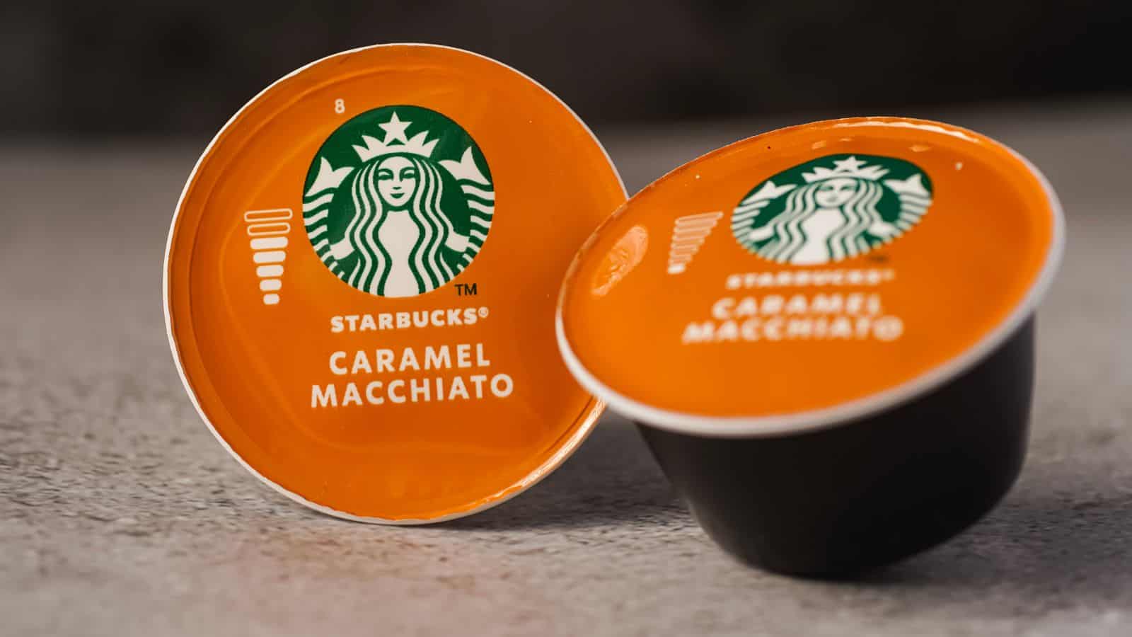 Two Starbucks Caramel Macchiato coffee pods are shown on a textured surface, one standing upright and the other tilted, both with orange lids featuring the Starbucks logo and product name.