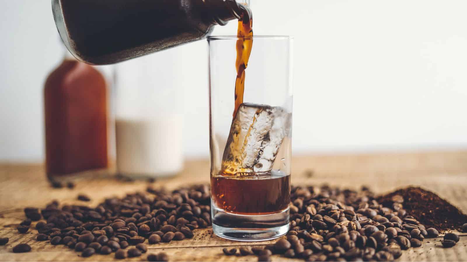 A glass with a large ice cube is being filled with dark coffee. Coffee beans are scattered on the table, and bottles containing milk and coffee are in the background.