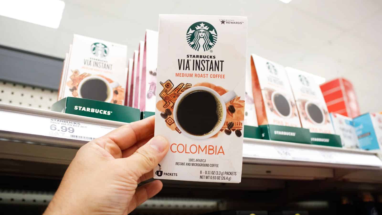A hand holds a box of Starbucks VIA Instant Colombia Medium Roast Coffee in front of a store shelf displaying other boxes of Starbucks coffee products. The price tag below reads $6.99.