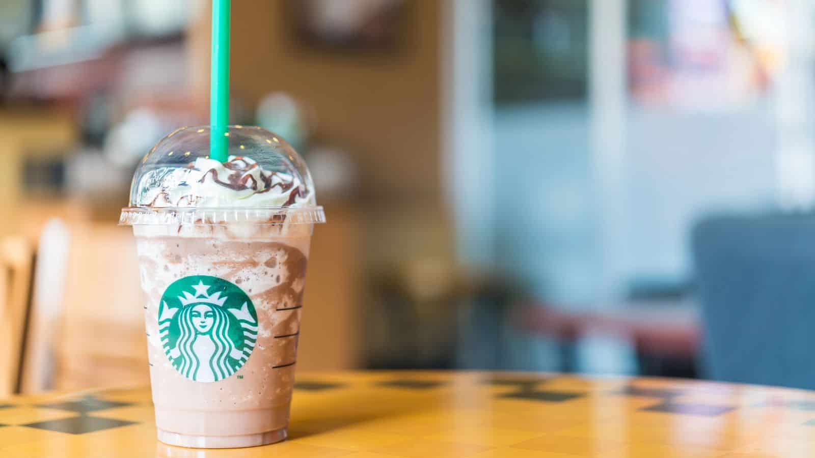 A Starbucks cold blended beverage with whipped cream and chocolate drizzle in a clear plastic cup with a green straw sits on a table. The background is blurred, showing an indoor café setting.